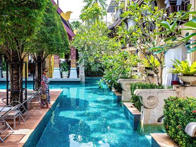 Schmaler blauer Pool zwischen Bäumen und Gebäuden mit Stühlen und dekorativen Pflanzen.