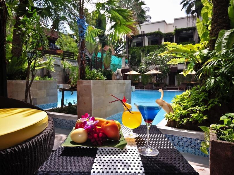 Bunte Früchte und Cocktails auf Tisch am Pool mit grünen Pflanzen und Sonnenschirmen im Hintergrund.