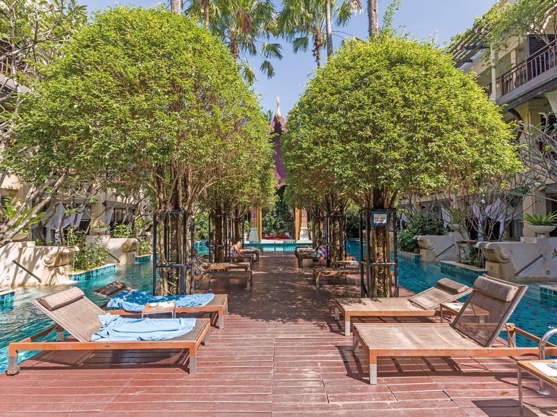 Poolbereich mit Holzsteg, Liegen und symmetrisch geschnittenen Bäumen unter blauem Himmel.