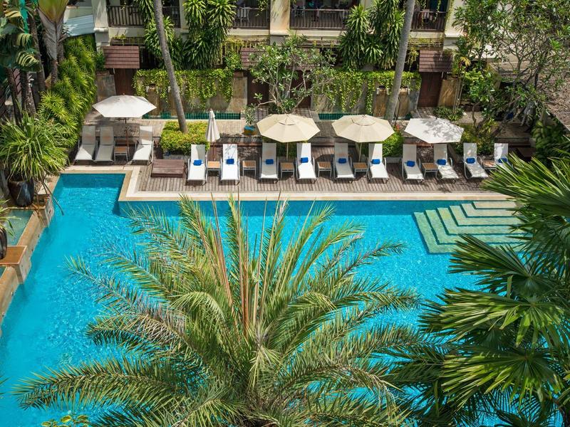 Blauer Pool mit weißen Liegestühlen und Sonnenschirmen, umgeben von Palmen und grünen Pflanzen.