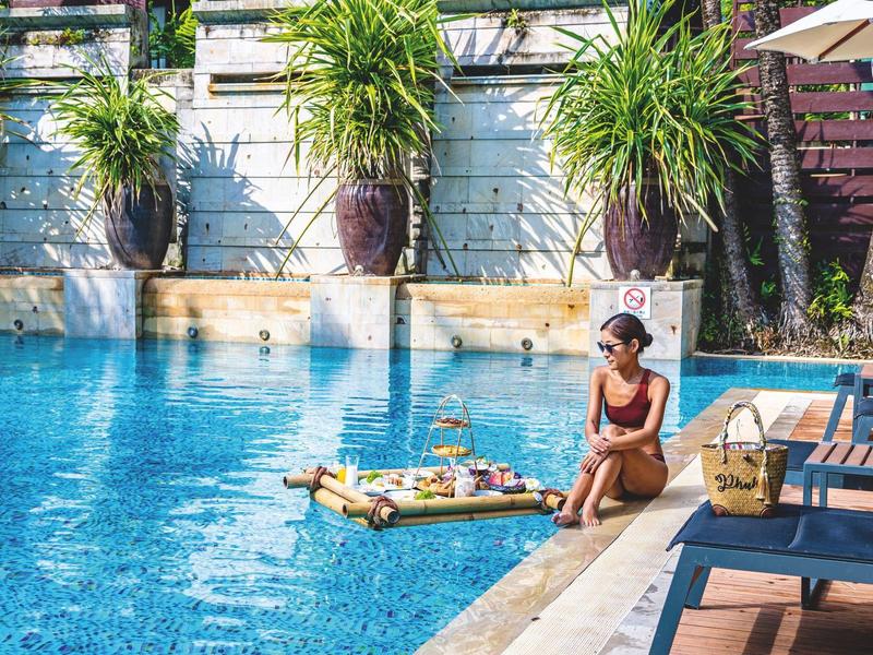 Frau sitzt am Poolrand, neben ihr schwimmt ein schwimmendes Frühstückstablett im blauen Wasser.