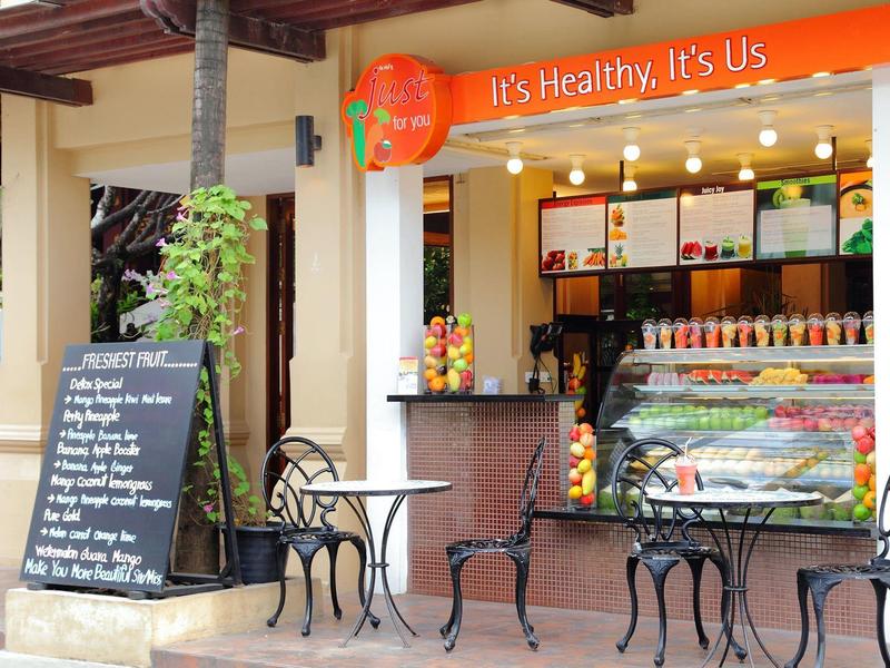 Offener Saftladen mit frischen Früchten, Eis, orangefarbenem Schriftzug und außen Metalltischen.