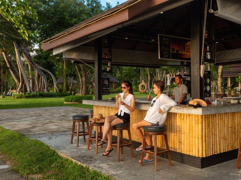 Zwei entspannte Personen sitzen an einer Strandbar mit Bambusdesign in tropischer Umgebung.