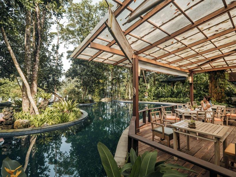 Überdachte Terrasse mit Tischen neben einem geschwungenen Pool in einer grünen, bewaldeten Umgebung.