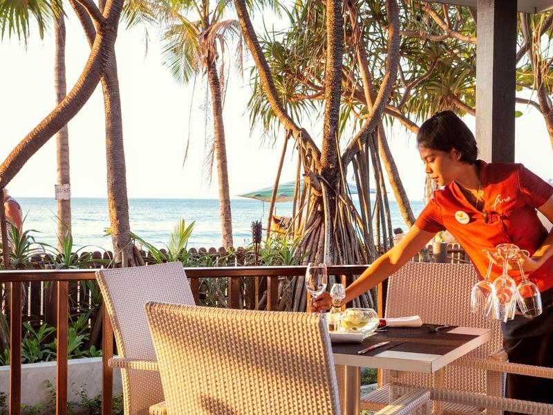 Ein Hotelangestellter bereitet auf einer Terrasse neben Palmen einen Tisch mit Getränken vor.