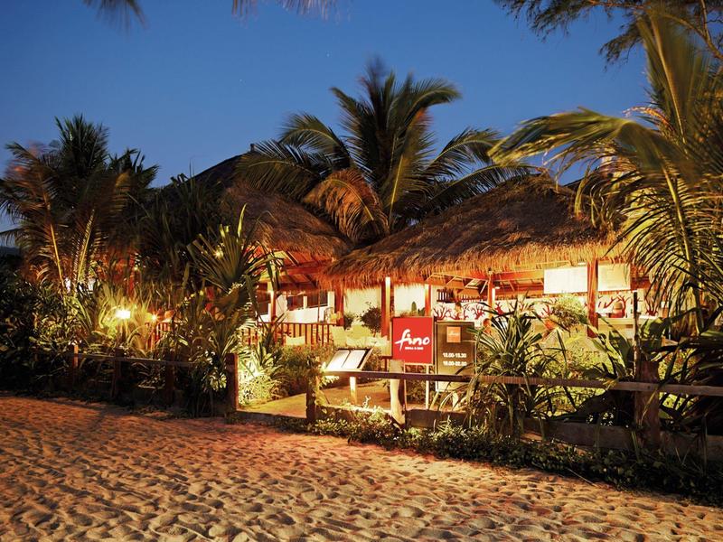 Beleuchtetes Strandrestaurant mit Strohdach und Palmen bei Dämmerung.