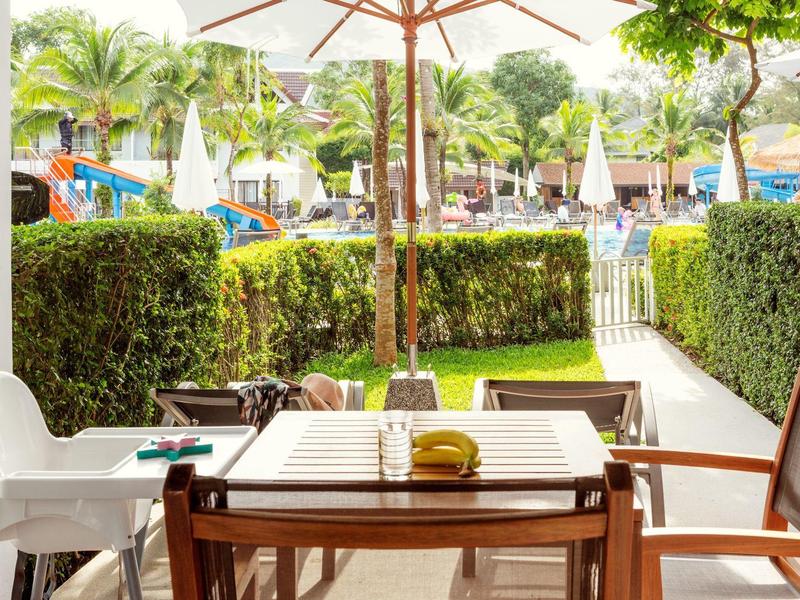 Blick von einer Terrasse mit Tisch, Stühlen und Sonnenschirm auf einen Poolbereich mit Palmen und Liegestühlen.