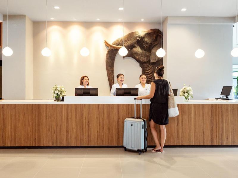 Hotelrezeption mit drei Mitarbeitern hinter dem Holztresen und einer Frau mit Koffer im Vordergrund.