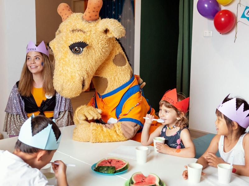 Kinder sitzen an einem Tisch mit Wassermelonenstücken, eine Person in einem Giraffenkostüm ist dabei.