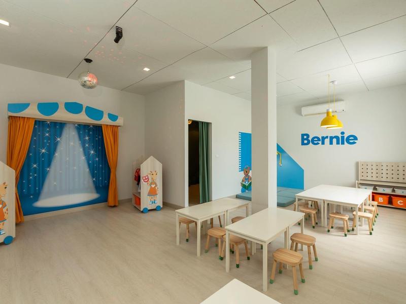 Heller, moderner Kinderspielraum mit Tischen, Stühlen und einer Bühne mit Vorhängen.