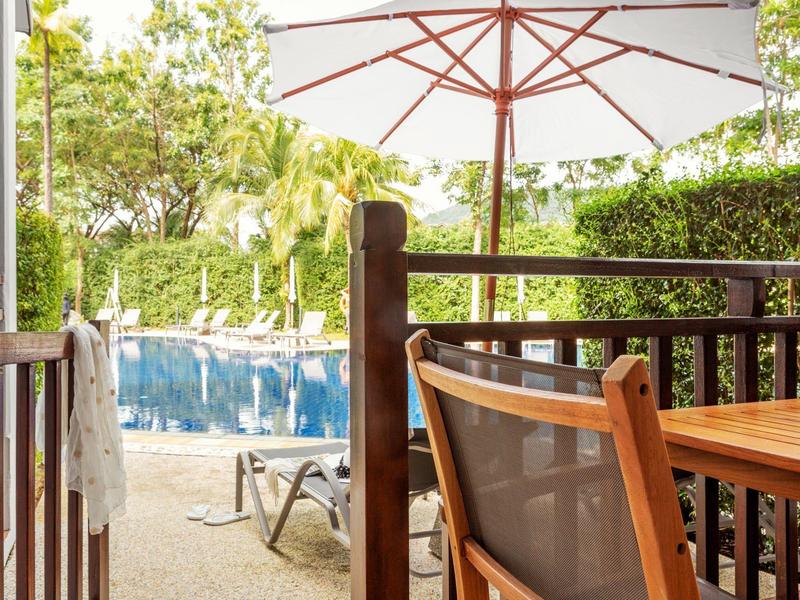 Blick von einer Terrasse mit Holzgeländer auf einen Pool und Liegestühle unter Bäumen.