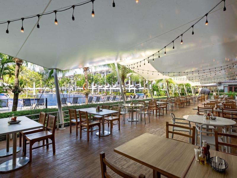 Überdachter Außenbereich eines Restaurants mit Holztischen und -stühlen neben einem Pool und tropischer Vegetation.