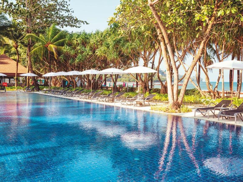 Großer Pool mit Sonnenliegen und weißen Sonnenschirmen neben tropischen Bäumen im Resort.
