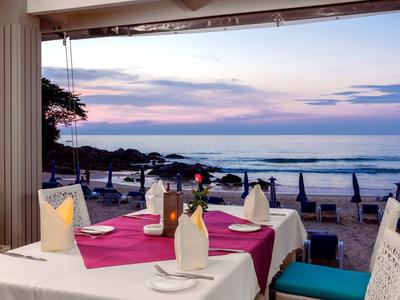 Strandrestaurant mit gedecktem Tisch, lila Tischdecke, weißen Stühlen und Blick auf das Meer bei Sonnenuntergang.