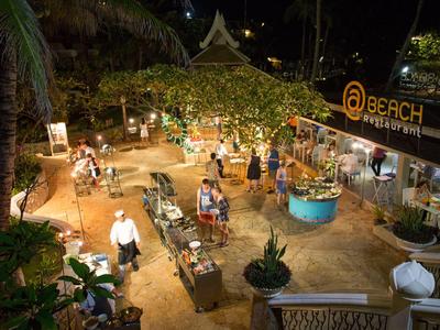 Strandrestaurant bei Nacht mit Tischen, Sitzgelegenheiten, Pflanzen und beleuchtetem Eingangsschild.