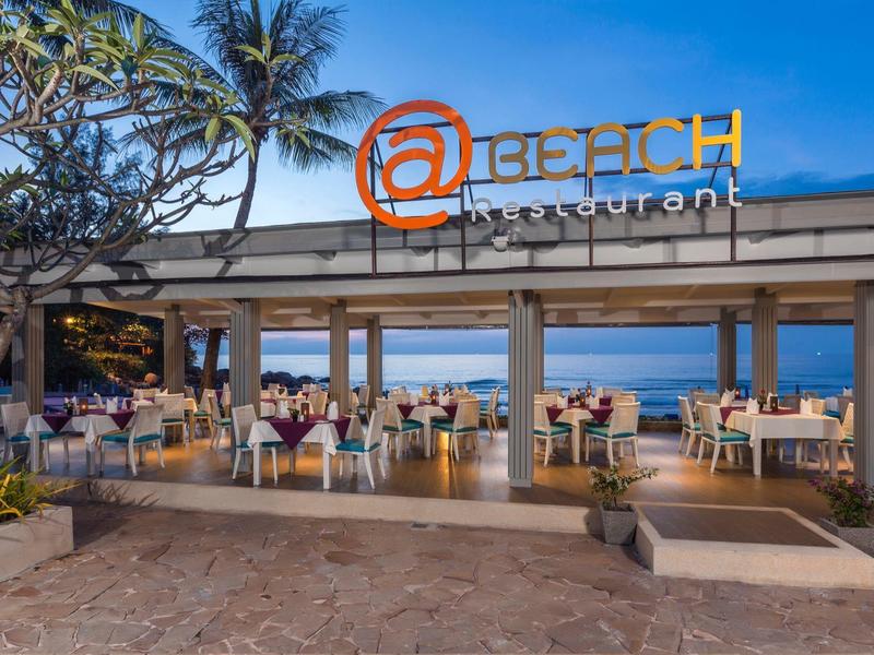 Offenes Strandrestaurant mit Tischen und Stühlen, Palmen und blauem Himmel im Hintergrund.