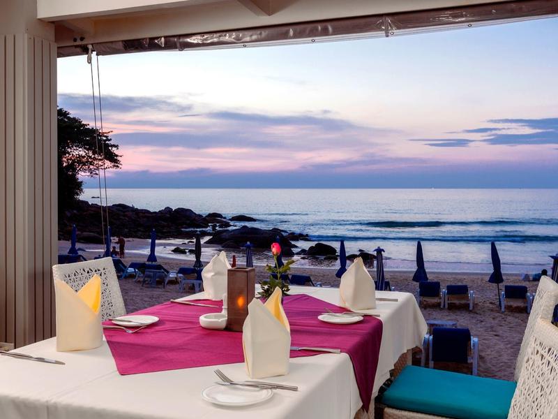 Strandrestaurant mit gedecktem Tisch, lila Tischdecke, weißen Stühlen und Blick auf das Meer bei Sonnenuntergang.