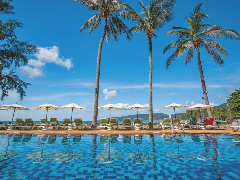 Blauer Pool mit Liegestühlen, Sonnenschirmen und hohen Palmen unter blauem Himmel.