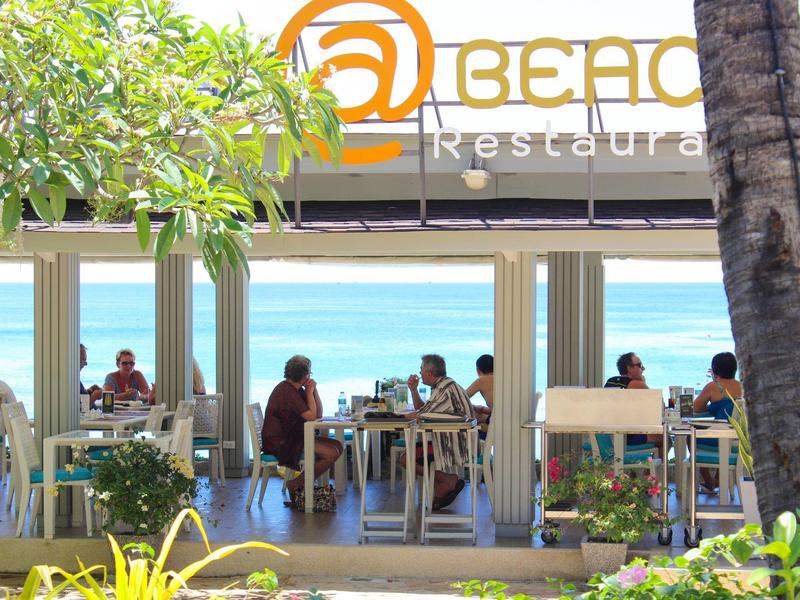 Restaurant am Strand mit Gästen, Palmen und Meerblick bei strahlendem Wetter.