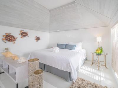 Hell eingerichtetes Schlafzimmer mit weißem Bett, grauen Kissen, Wanddeko und seitlichem Beistelltisch.