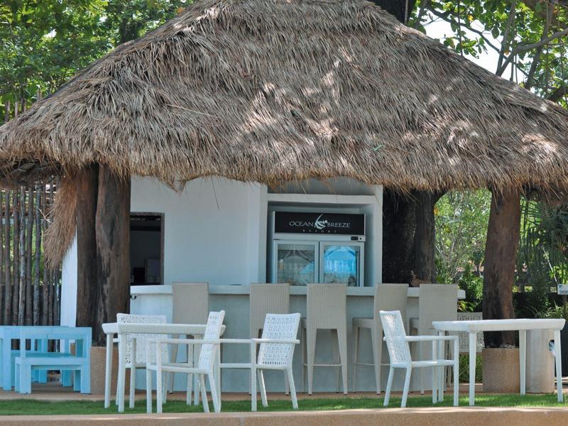 Offene Bar mit Strohdach und weißen Stühlen in einer tropischen Umgebung.
