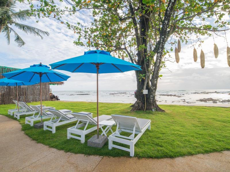 Weiße Sonnenliegen mit blauen Sonnenschirmen auf grünem Rasen neben einem Baum und Blick auf das Meer.