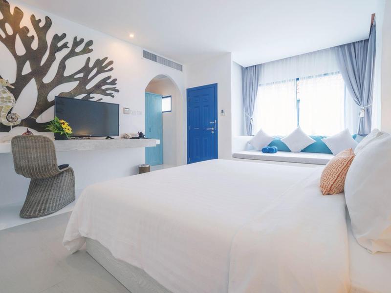 Helles Hotelzimmer mit großem Bett, blauer Tür und einem Fensterplatz mit Kissen.