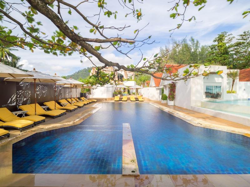 Schwimmbad mit blauen Fliesen, gelben Liegestühlen und schattigen Bereichen unter Bäumen.