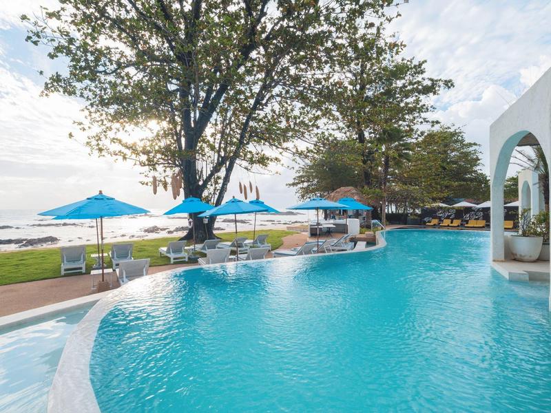 Großer Pool mit blauen Sonnenschirmen und Liegestühlen neben einem Baum und Meerblick