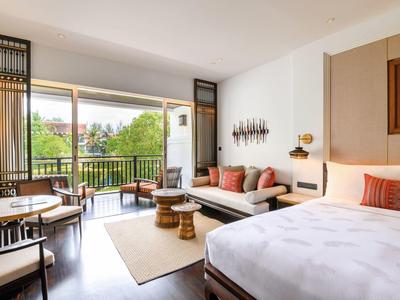 Helles Hotelzimmer mit großem Bett, Sitzbereich und Balkon mit Blick auf grüne Bäume.