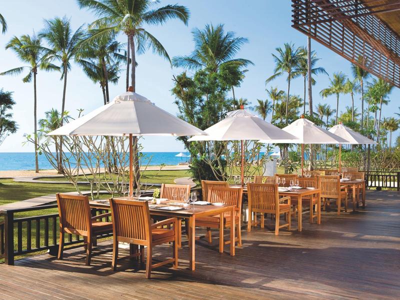 Terrasse mit Holztischen und Stühlen, Sonnenschirmen und Palmen im Hintergrund am Meer.