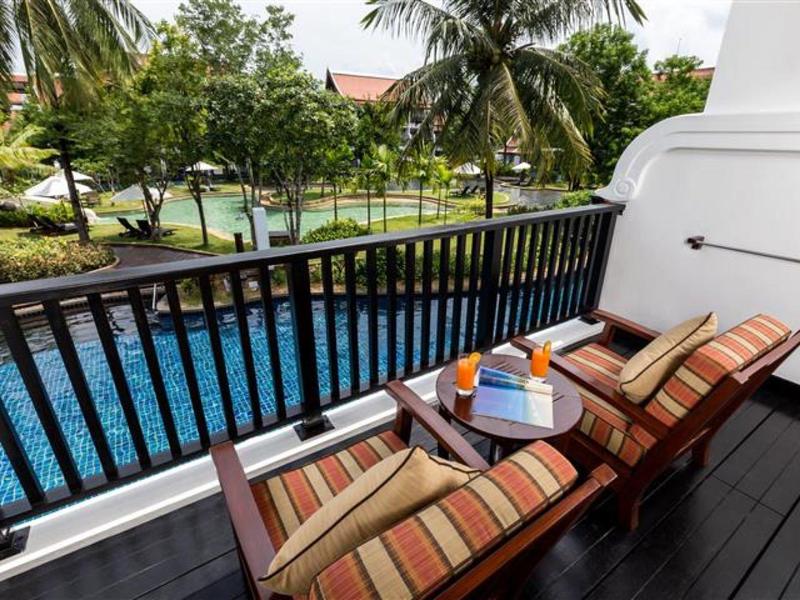 Balkon mit zwei Liegestühlen, kleinem Tisch und Blick auf Pool und Palmen in tropischer Umgebung.