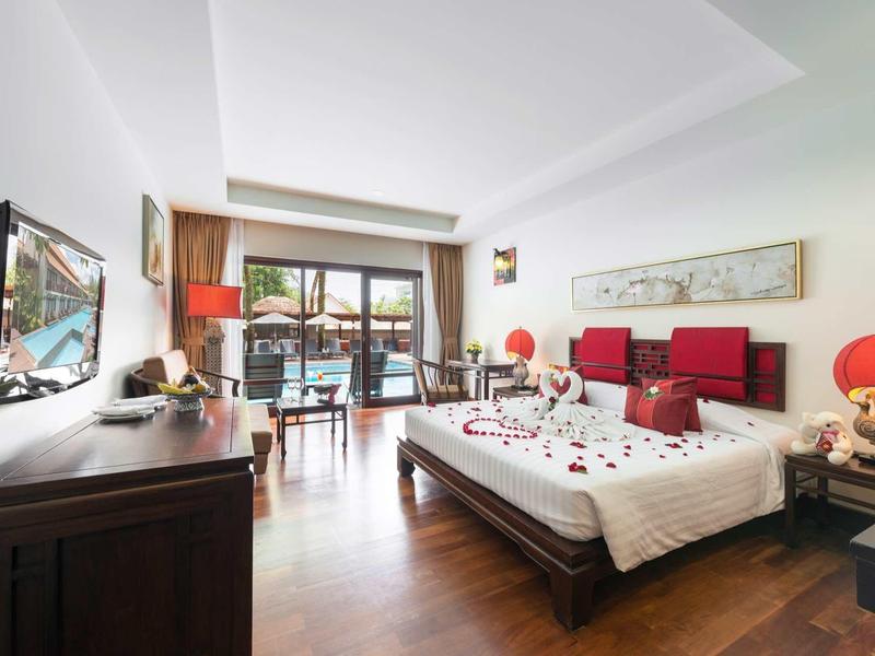 Helles Wohnzimmer mit Holzboden, rotem Sofa, TV, großem Bett und Balkontür mit Blick nach draußen.