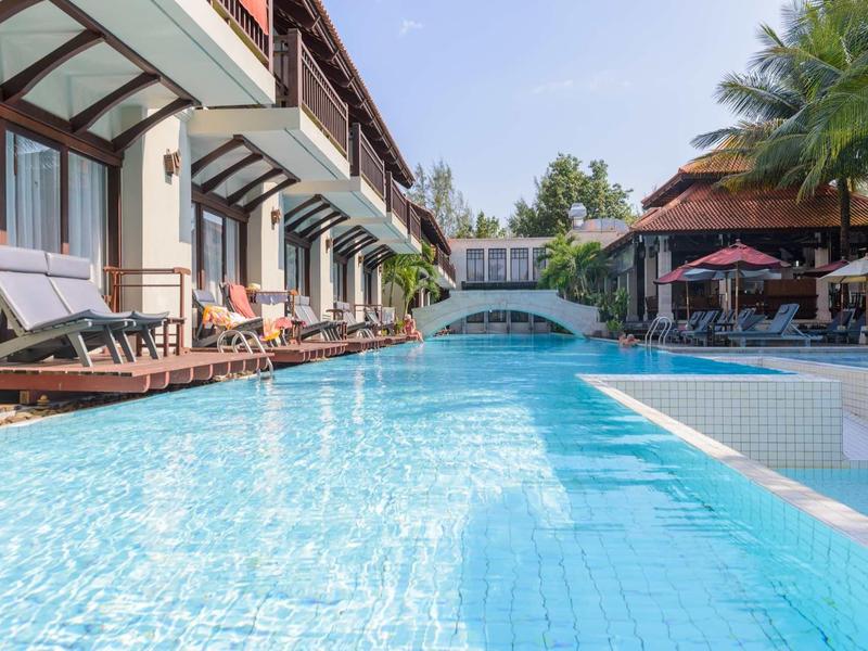 Großer, klarer Pool mit Sonnenliegen neben modernen Gebäuden und einer kleinen Brücke im Hintergrund.