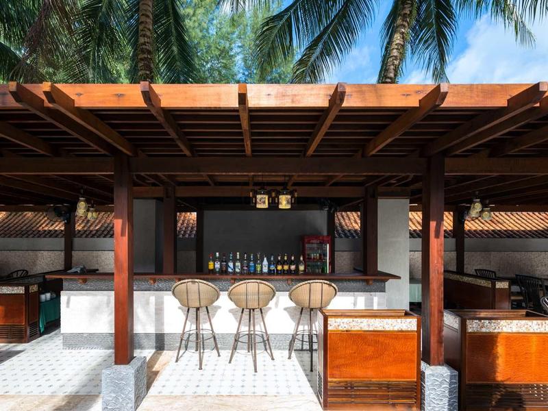 Offene Bar mit Holzstruktur, Hockern und tropischen Pflanzen im Hintergrund.