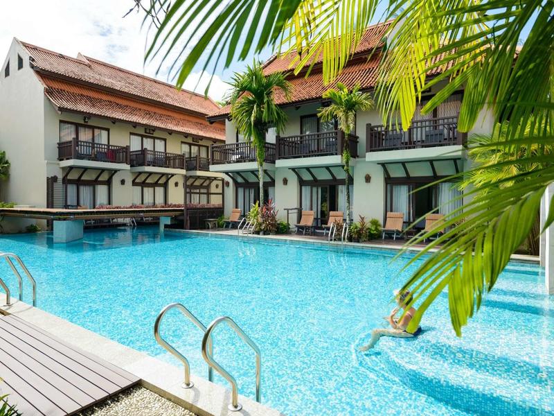 Großer blauer Pool vor einem zweistöckigen Hotelgebäude mit viel Glas und Palmen im Vordergrund.
