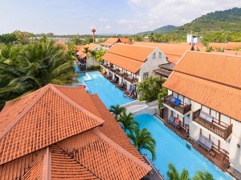 Hotelanlage mit roten Ziegeldächern, einem blauen Pool und Palmen in sonniger Landschaft.
