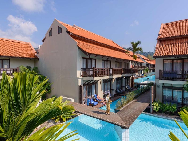 Hotel mit roten Dächern, Holzterrassen und blauem Pool unter blauem Himmel.