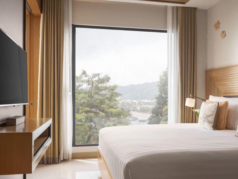 Camera d'hotel moderna con letto grande, TV a parete e vista su alberi e montagne.