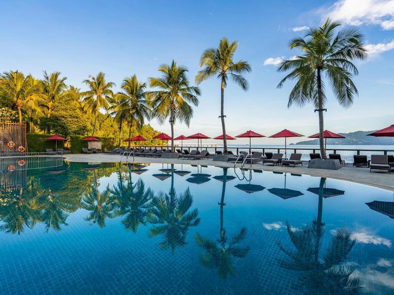 Una piscina tranquilla con palme e lettini sotto ombrelloni rossi vicino alla spiaggia.