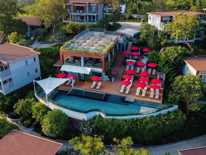 Vista aerea di un hotel con piscina, terrazza solarium e ombrelloni rossi in un contesto verde.