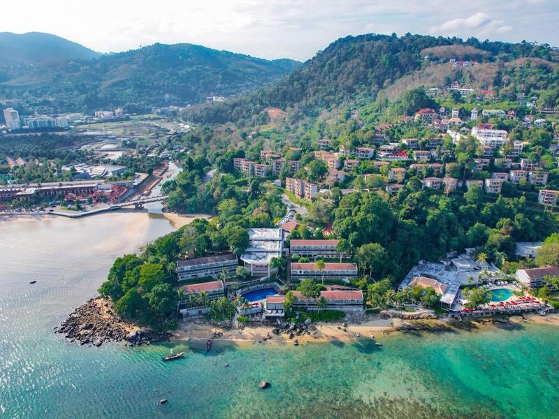 Veduta aerea di una città costiera con colline, spiaggia e acqua limpida sotto il sole.