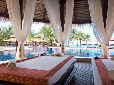 Offene Massagekabine mit weißen Vorhängen, zwei Massageliegen und Blick auf Pool und Palmen.