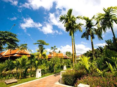 Tropischer Garten mit Palmen, bunten Pflanzen und einem gepflasterten Weg unter blauem Himmel.