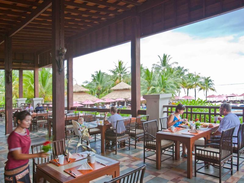 Überdachte Terrasse eines Restaurants mit Holzmöbeln, Gästen und Blick auf Palmen und Wasser.