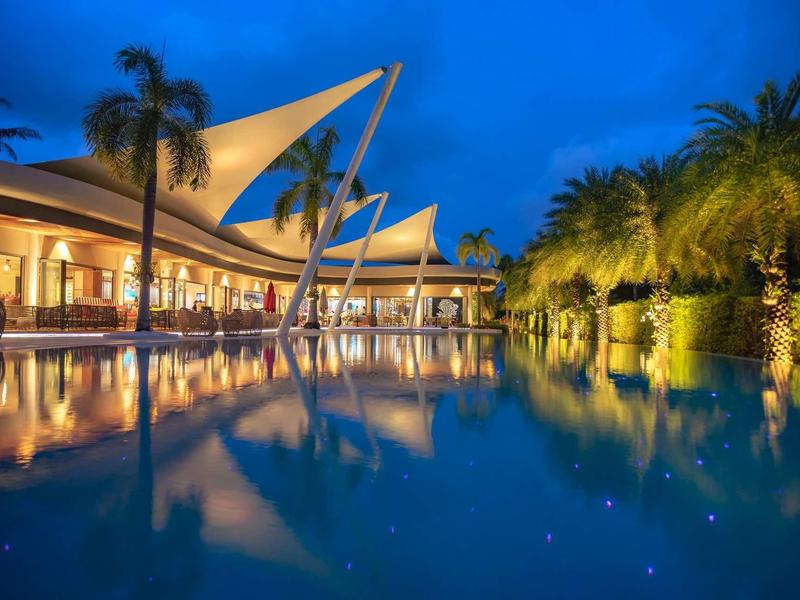 Piscina moderna dell'hotel con palme e tetti a vela illuminati di notte.