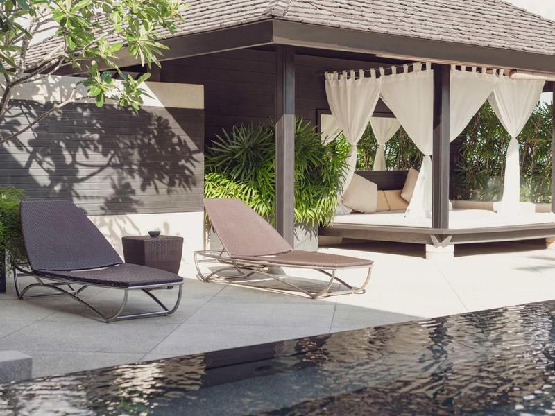 Area piscina moderna con lettini e lounge esterno coperto circondato da piante.