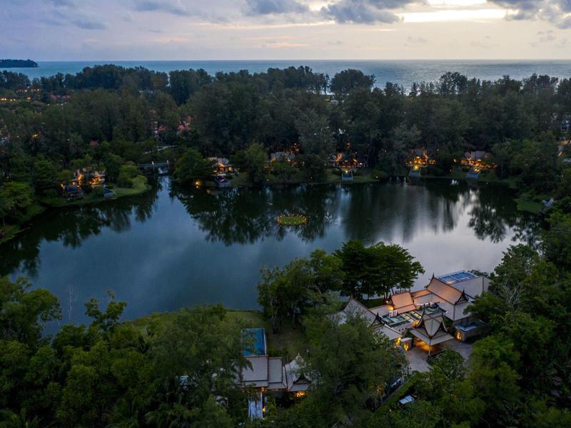 Runder See mit Häusern und vielen Bäumen, dahinter Meer unter bewölktem Himmel bei Dämmerung.