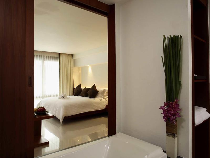 Moderner Badebereich mit weißer Badewanne, Sicht auf Schlafzimmer mit Bett und Kissen, Pflanzen-Deko.