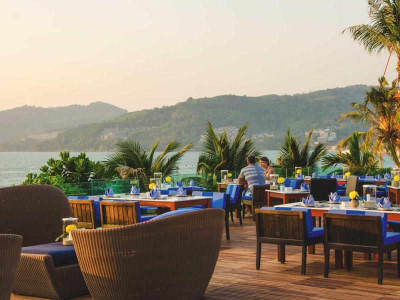 Terrassenrestaurant mit Holzboden, blauen Tischdecken und Palmen vor Meer und Bergen im Sonnenuntergang.